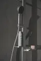 Handbrause von Ideal Standard, montiert an einer Duschstange, mit einem Halter für Shampoo und einem kleinen Ablagefach. Wand ist in einem grauen Schattenton gehalten.